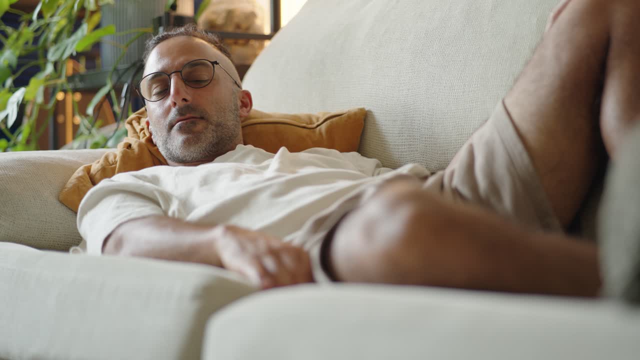 Relaxed middle aged man wearing reading glasses lies comfortably on the sofa, smiling warmly at the camera in a casual living room