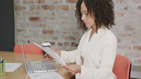 busy biracial businesswoman at desk reading document and using laptop at office, in slow motion