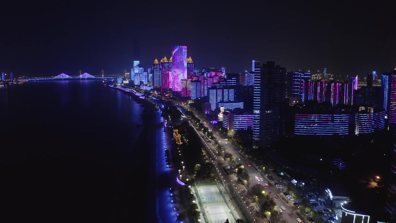 Panoramic night aerial view of illuminated modern skyscrapers along the Yangtze River in Wuhan, Hubei, China, displaying vibrant LED light shows, coastal road traffic, and distant lit bridges.