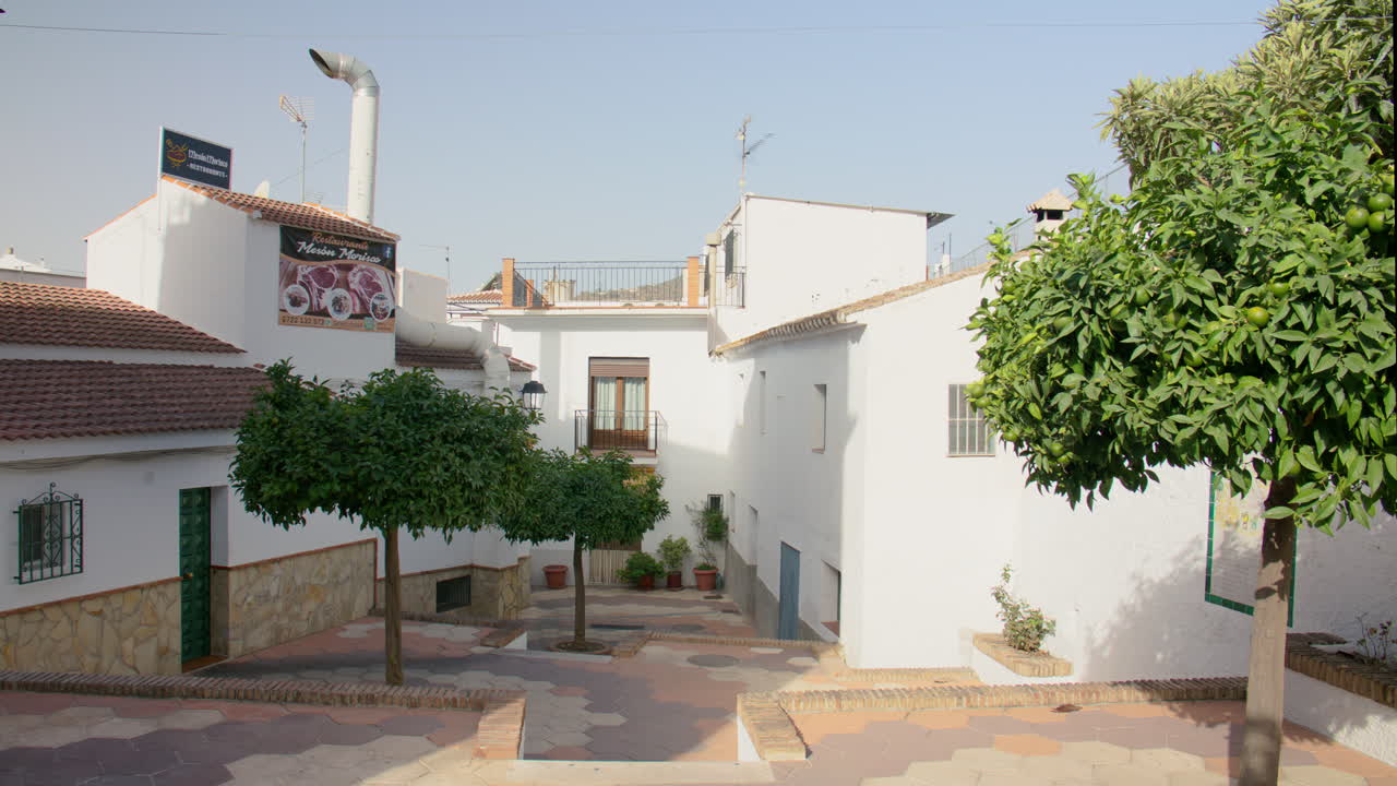 Peaceful morning light bathes a quiet alley in a quaint Spanish town, with white walls, tile roofs, and orange trees