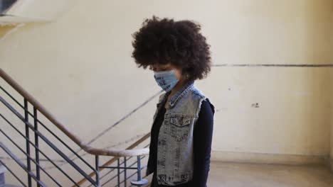 Portrait-of-african-american-woman-wearing-protest-face-mask-standing-near-the-stairs