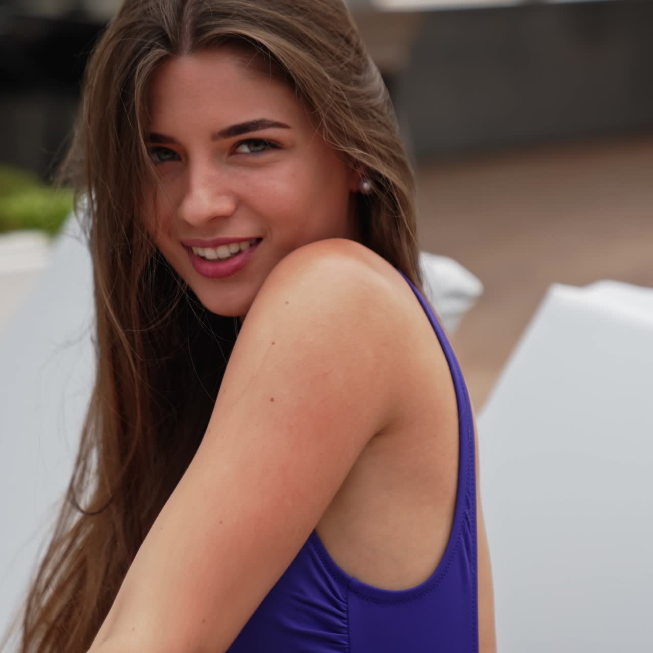 Lovely young lady touching her long beautiful dark hair. Relaxed girl leans back sitting on a deckchair in purple swimsuit