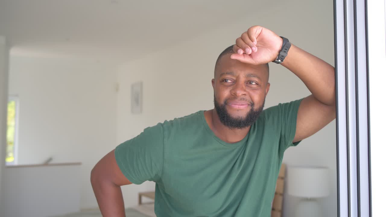Man wearing green tee and watch leaning on sliding door in bedroom, noticing balcony, smiling