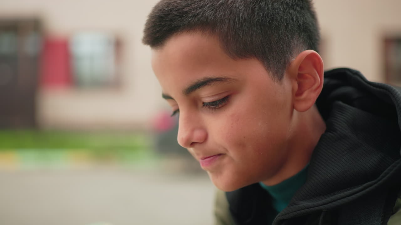 Close up side view of young kid concentrating on study with serious expression, blurred background featuring building and parked car showing focus, learning habit