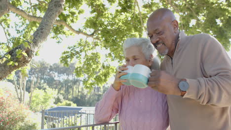 Happy-senior-biracial-couple-embracing-and-drinking-coffee-at-balcony-at-home,-slow-motion