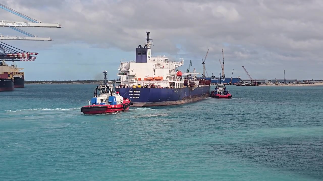 Plano general de dos remolcadores arrastrando y guiando el enorme buque de carga BW Wren a través de aguas azules en una concurrida terminal portuaria comercial
