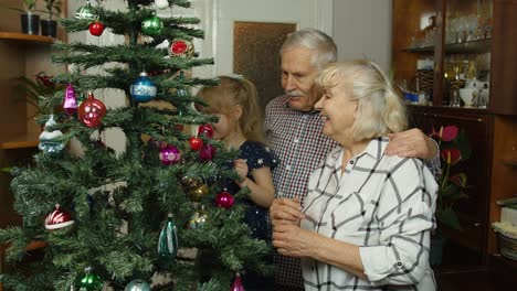 Child-girl-with-senior-grandparents-family-decorating-artificial-Christmas-tree,-New-Year-holidays