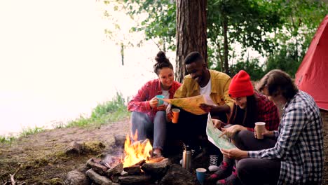 cinemagraph loop - group of young men and women are looking at paper maps sitting around fire in forest with drinks. map is moving, tent and trees are visible.