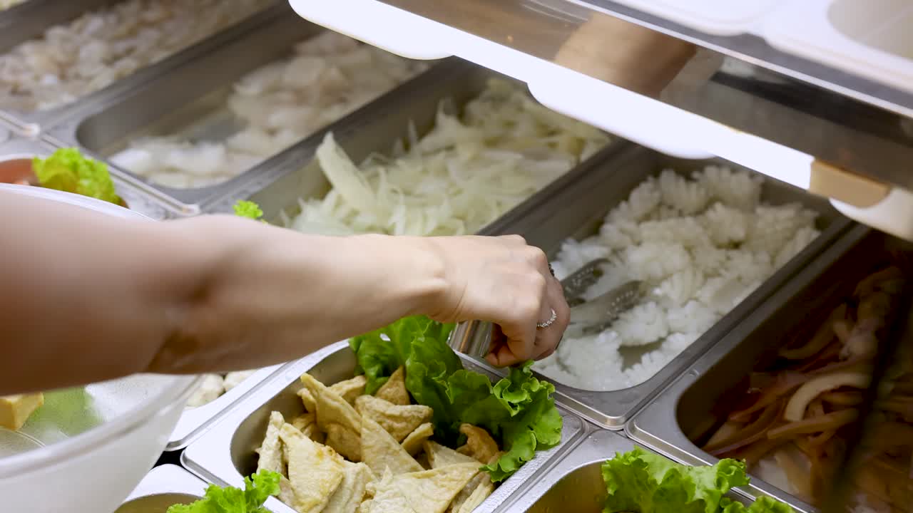Person uses tongs to pick tofu and vegetables from well-lit buffet counter, overhead view