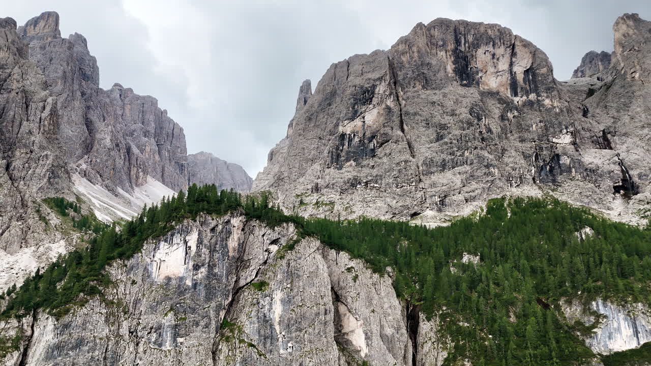 Luftfoto med drone af en massiv lodret kalkstensklippe, der rejser sig over tæt alpeskov. Den imponerende klippevæg dominerer Dolomitternes bjergrige landskab.