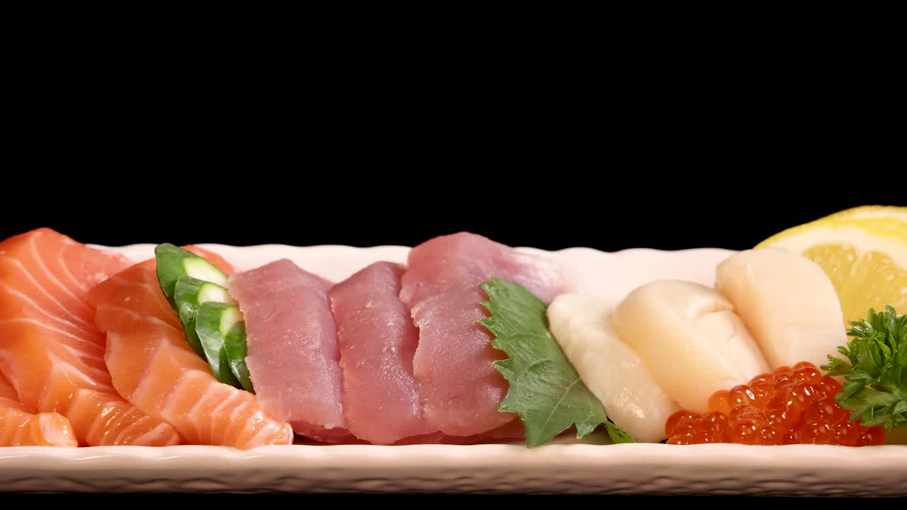 A rectangular platter of assorted sashimi and sushi, including salmon, tuna, scallop, and roe, rotates smoothly under bright studio lighting against a black background