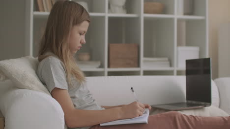 teen girl is doing homework in room of flat after school writing in exercise book sitting on couch e-learning for school children