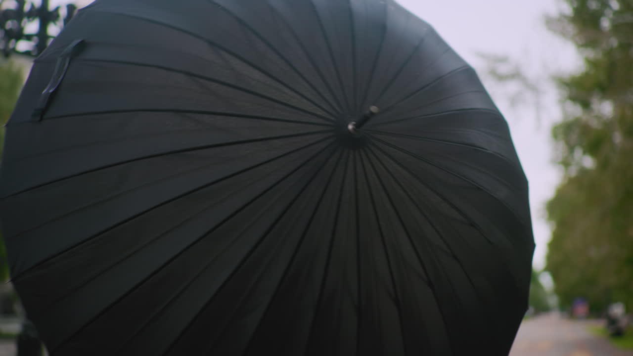 Person opening a black umbrella on city street during cloudy day with blurred background of trees buildings cars and lamppost showing urban lifestyle mood outdoor weather and seasonal atmosphere