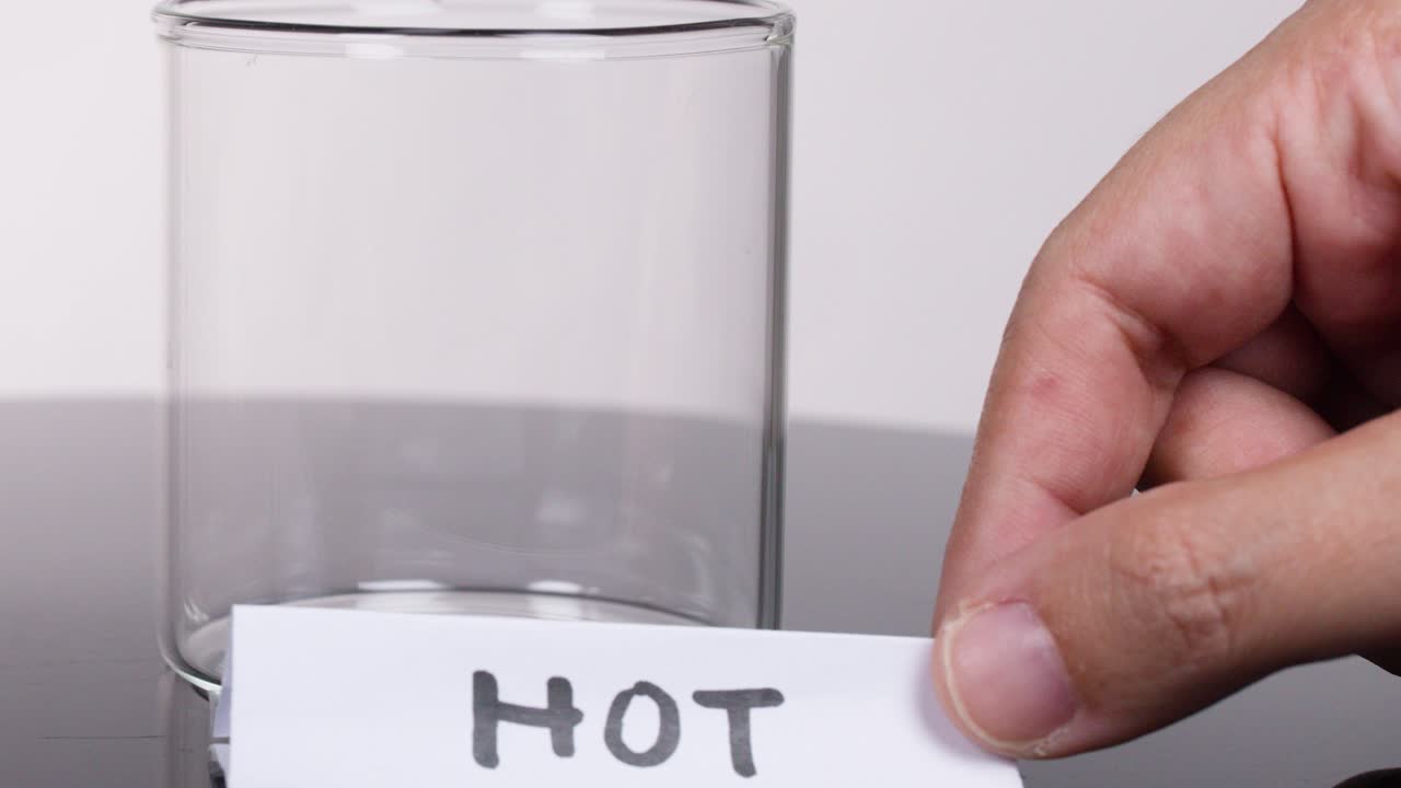 Hand places handwritten 'HOT' paper label next to clear glass beaker on laboratory table