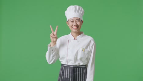 Asian Woman Chef Smiling And Showing Peace Gesture To Camera While Standing In The Green Screen Background Studio