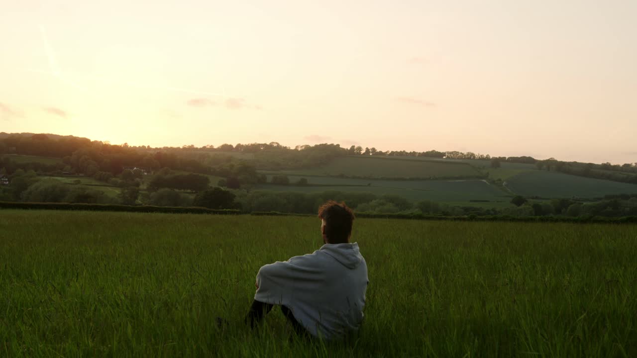 A man sits alone in a quiet field watching the sunset over distant hills