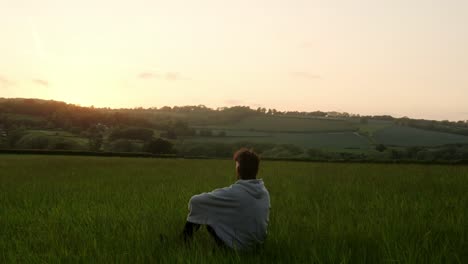 a man sits alone in a quiet field watching the sunset over distant hills