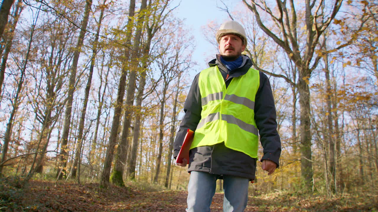 male engineer in safety vest and hard hat walking in the woods alone while carrying clipboard, handheld tracking shot