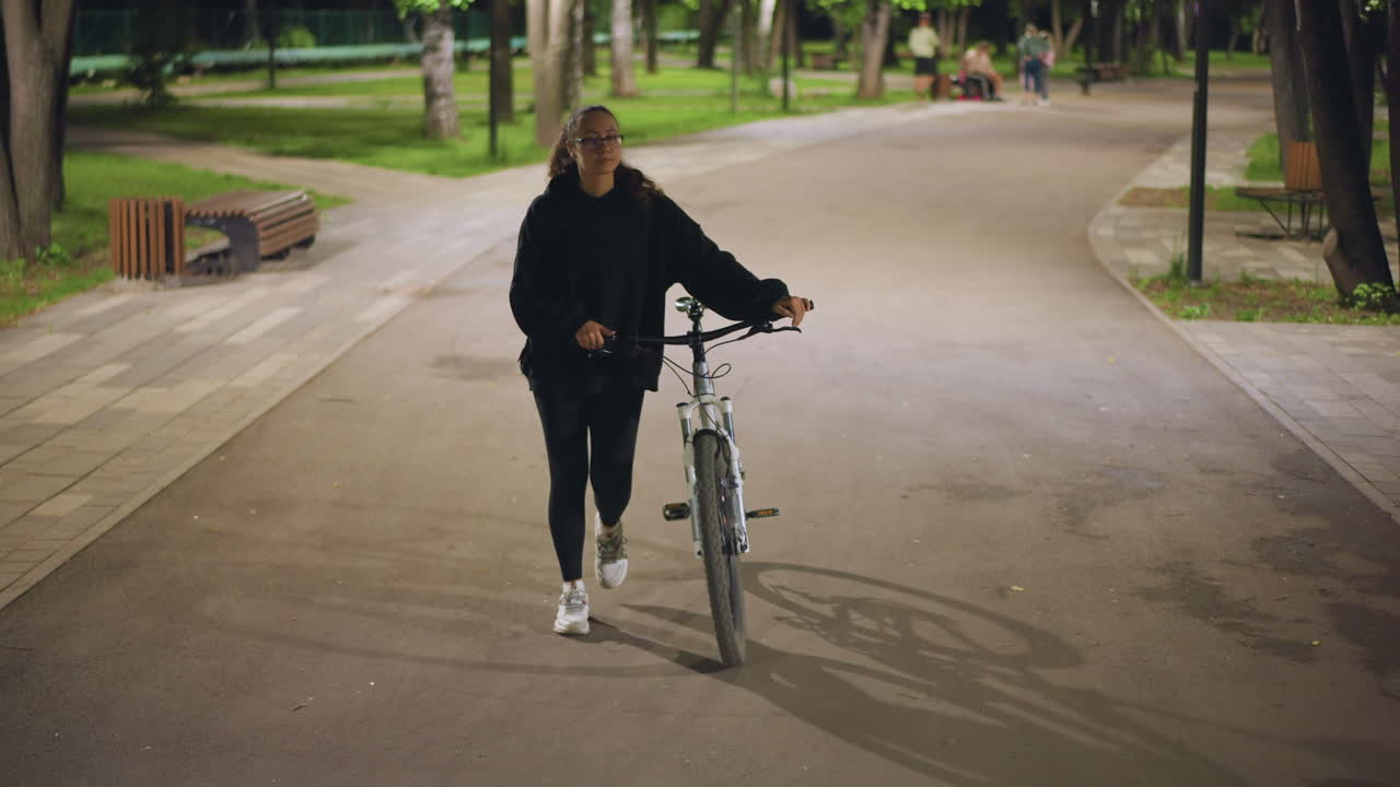 Caucasian Woman Walking Bicycle On Path, Frontal View In Illuminated Park With Benches And Trees, Wearing Hoodie And Sneakers While Calmly Pushing Bike Along Paved Walkway, Steady Pace And Relaxed