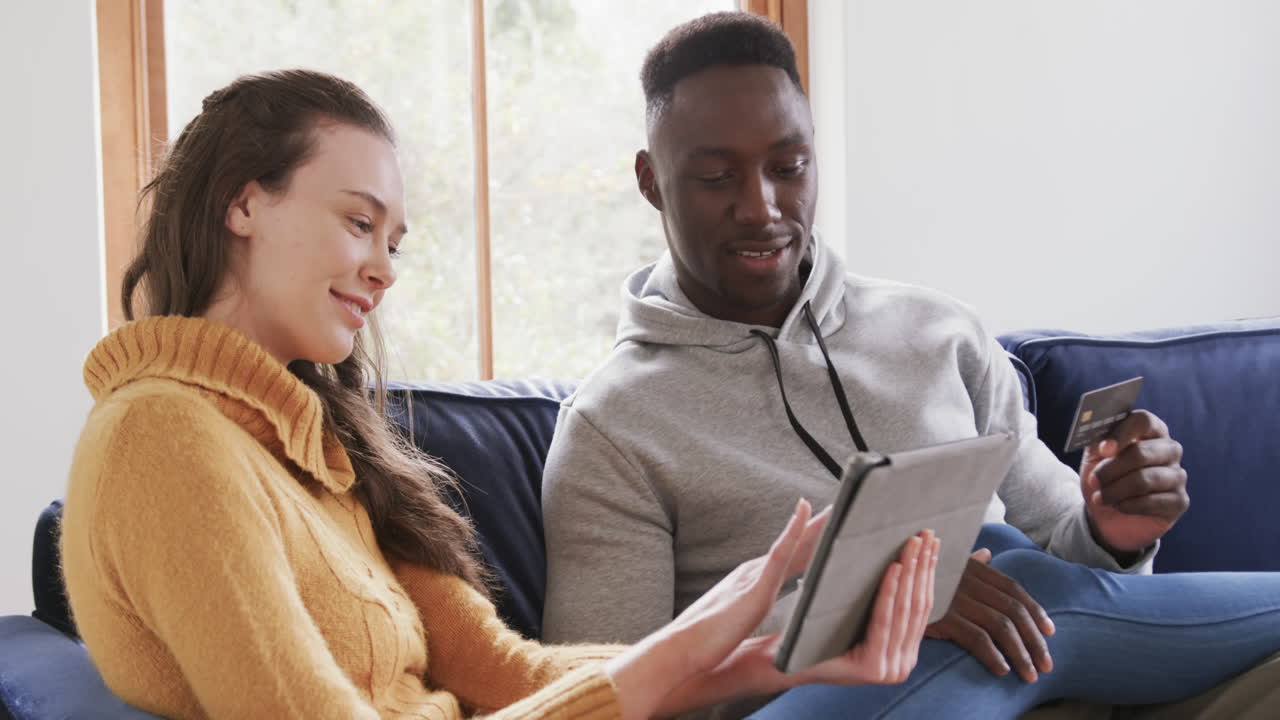 Happy diverse couple sitting on sofa, using tablet and credit card in home,copy space
