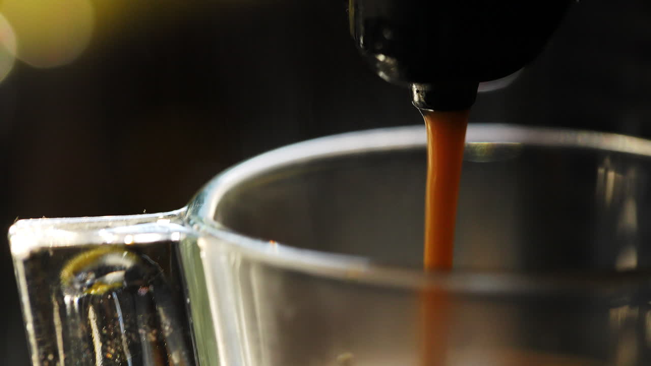 Freshly brewed coffee cascading from professional espresso machine into transparent glass cup, creating mesmerizing ripples across dark liquid surface