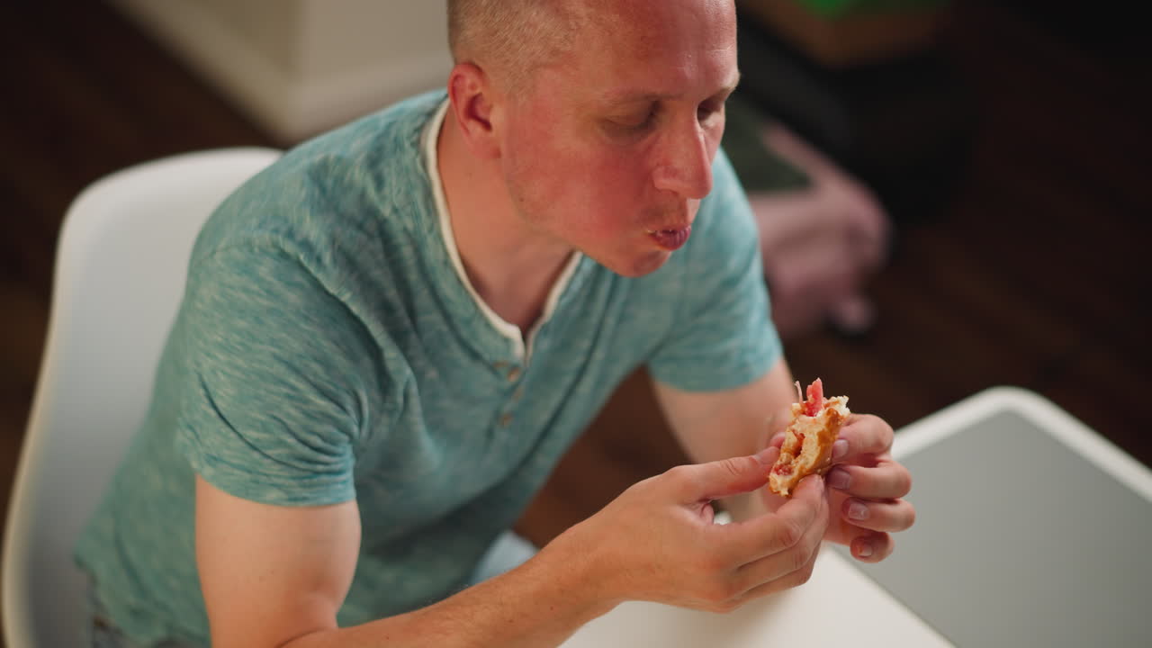 Aerial view of adult man wearing teal shirt eating snack at white table with blur background featuring unclear item, enjoying his meal with both hands