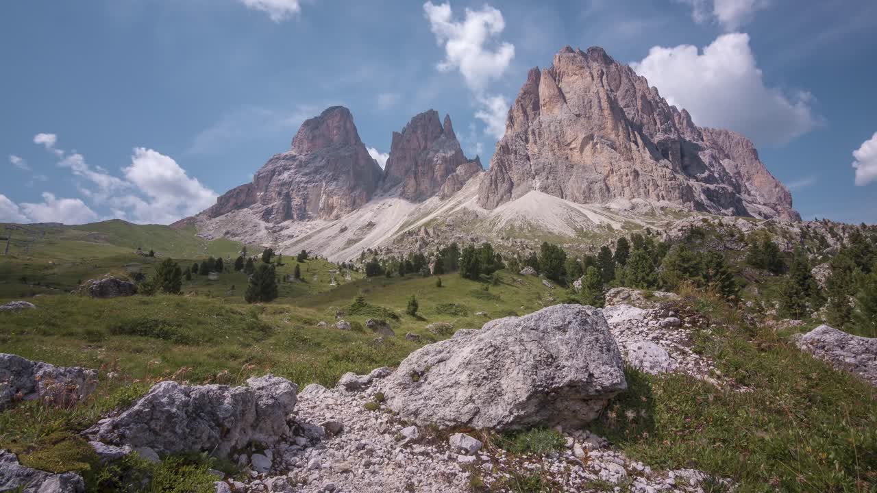 Timelapse of the stunning Langkofel in the Dolomites with clouds gracefully moving across the frame. Sunlight filters through, revealing a vibrant blue sky behind the clouds.