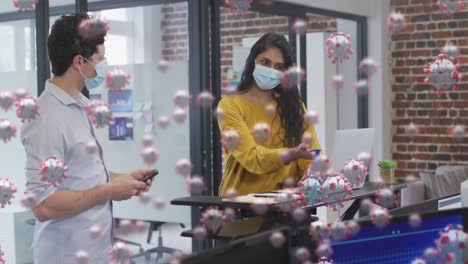 animation of covid 19 cells floating over man and woman wearing face masks in office, discussing