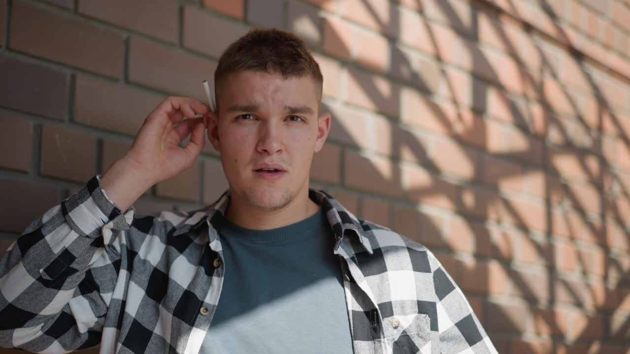 young man in black white check shirt returns cigarette to ear while gazing into distance from metal staircase landing against brick wall backdrop with dappled sunlight and lush greenery beyond