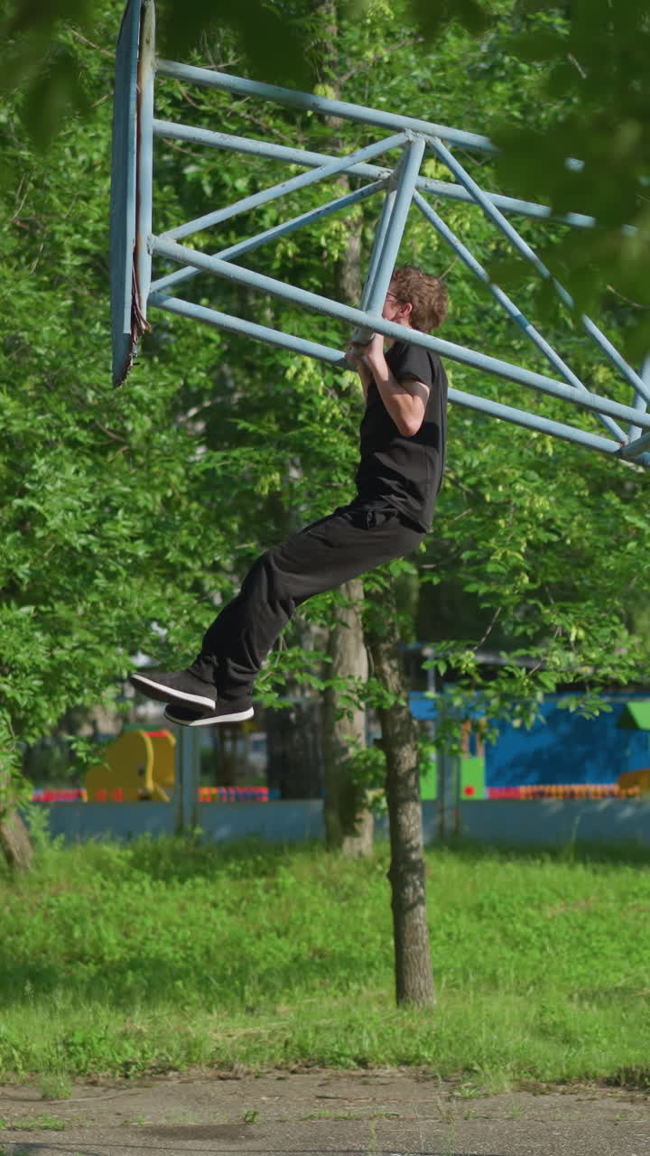 A young boy dressed in a black outfit performs multiple pull-ups, gradually lifting his head over the bar on outdoor equipment, with lush green trees and fence in the background