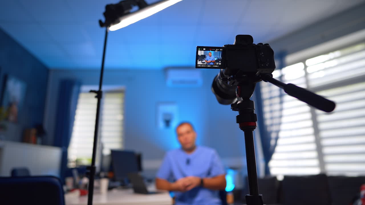 Light and camera directed on the man at blurred backdrop. Male in blue uniform talks gesturing in front of camera for his blog. Selective focus