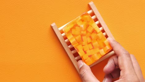 primer plano de una persona sosteniendo una barra de jabón hecha a mano de color naranja en un plato de jabón de madera