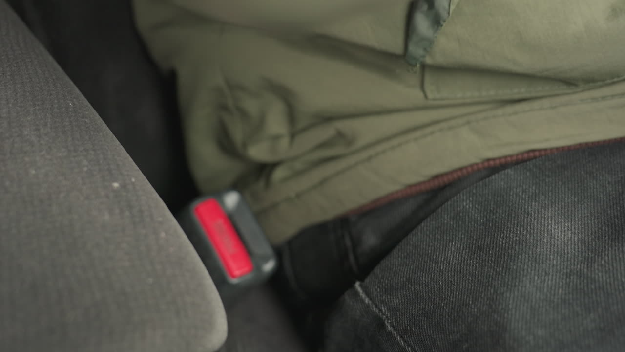 close up of person in black jeans and green jacket removing seatbelt from buckle with firm grip inside car, showing moment of disengagement from restraint system with focus on hand and seat texture