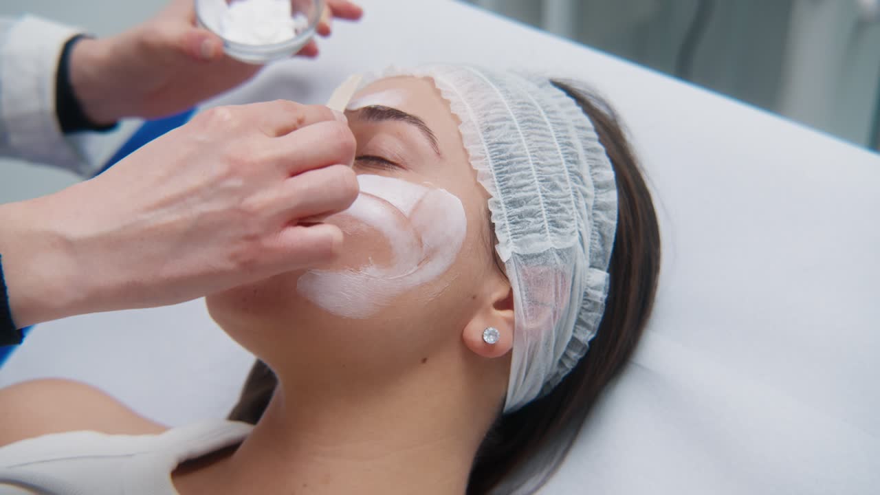 Esteticista aplica una crema facial blanca y espesa en la mejilla de una mujer usando una espátula durante una sesión de cuidado de la piel en un spa