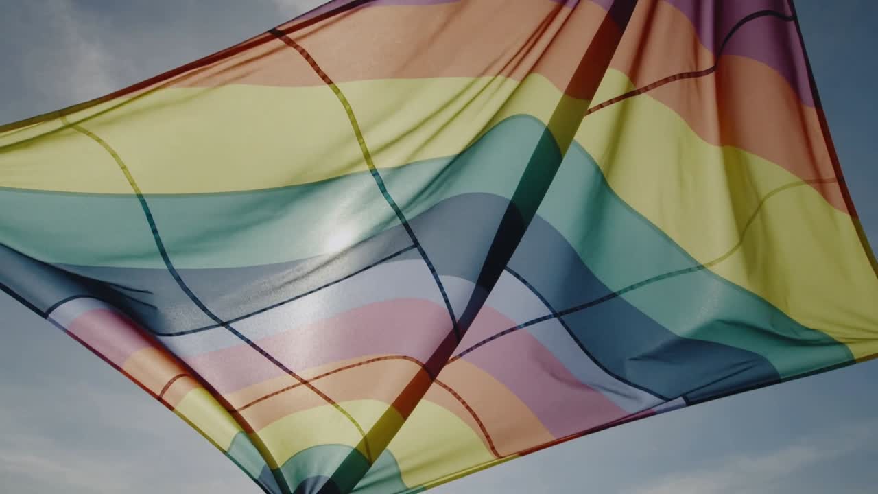 Grande bandiera arcobaleno che ondeggia dolcemente per una leggera brezza contro un cielo aperto, con la luce del sole che evidenzia le cuciture.