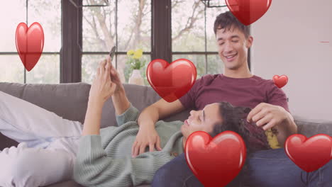 animation of red hearts over happy diverse gay male couple relaxing on couch using smartphone