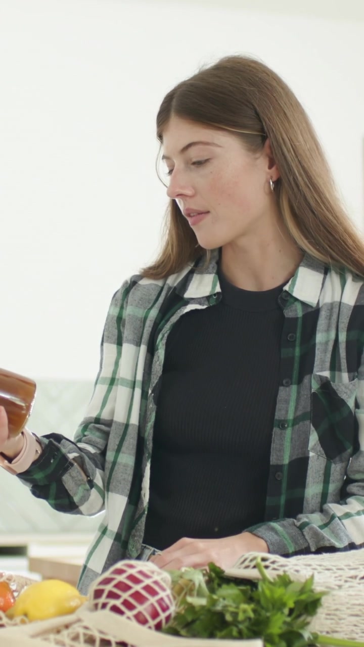 Vertical video: Reaching woman wearing plaid unpacking net bag in kitchen, checking amber jar label