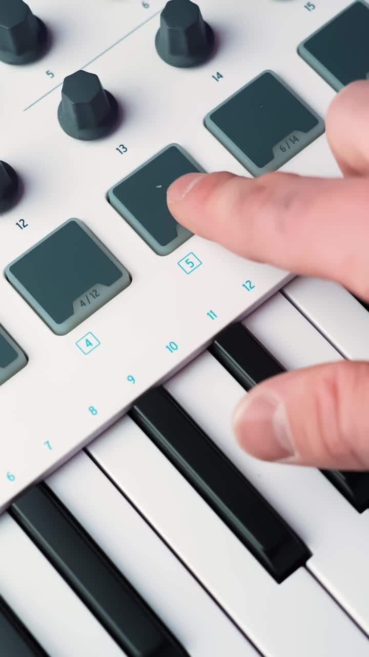 Close-up tangan mengetuk bantalan drum pada pengontrol keyboard MIDI, memicu ketukan dan akor dalam pengaturan studio rumah yang bersih dengan kedalaman bidang dangkal, vertikal.