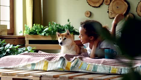 young woman is stroking lovely puppy and talking to it expressing love while they are lying on bed in modern flat together resting. loving animals and youth concept.