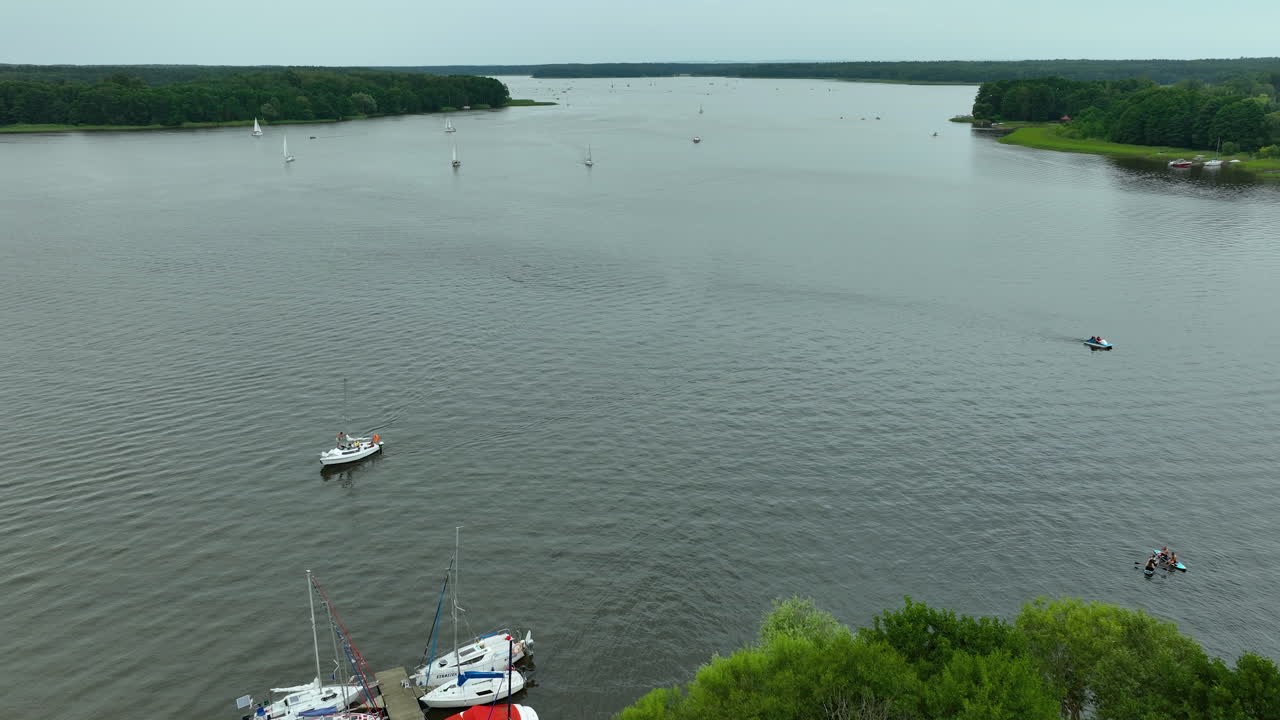Aerial view of a small pier extending into Jeziorak lake, with sailboats moored nearby and trees surrounding the shoreline, creating a tranquil and scenic atmosphere