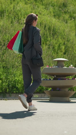 mujer con ropa gris caminando con bolsas de compras de colores detrás de ella, paseando al lado de una colina en un día soleado, el fondo presenta vegetación y un banco bajo la luz del sol brillante