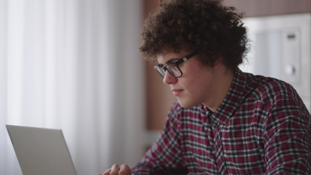A Curly man with a serious look works at a laptop sitting in a modern kitchen. Young man freelancer student using laptop studying online working from home in internet