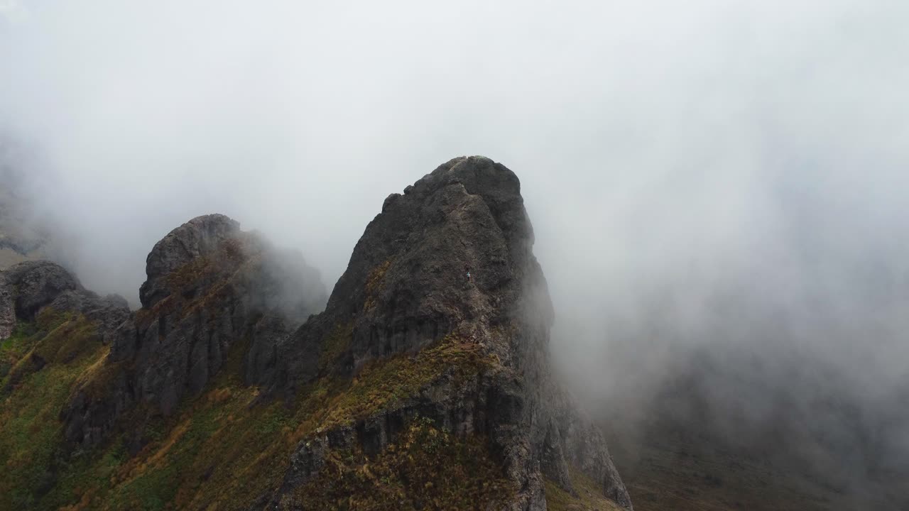 Aerial dolly-in and tilt-down shot capturing two mountaineers scaling a steep, exposed peak at Cerro Puntas, Ecuador. Surrounded by mist, they navigate the extreme ascent on the rugged cliffside.