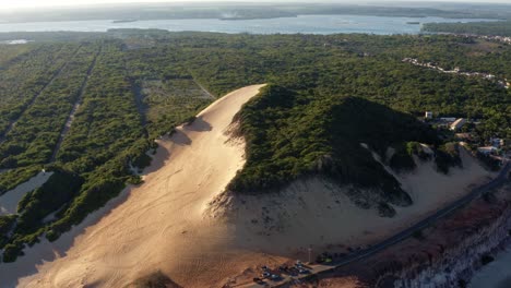 Toma-Aérea-Rotativa-De-Drones-Del-Famoso-Destino-Turístico-Cacimbinhas-Duna-De-Arena-Con-Turistas-Sandboarding-Cerca-De-Pipa,-Brasil-En-Rio-Grande-Do-Norte-Con-La-Laguna-Guaraíras-En-El-Fondo