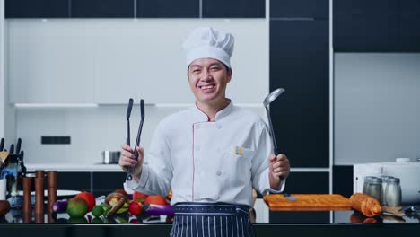 asian man chef smiling and showing tongs and ladle to camera while standing in home kitchen