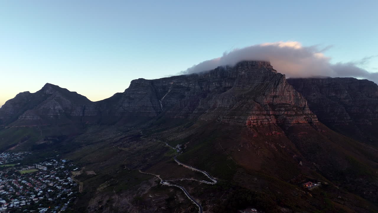 Cinematic aerial view of Table Mountain in Cape Town, South Africa, dramatic rocky cliffs with coastal cityscape at sunset, clouds rolling over peaks and golden evening light