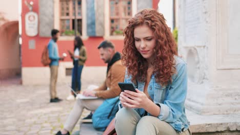 mujer pelirroja usando un teléfono inteligente mientras se sienta en el suelo en la calle