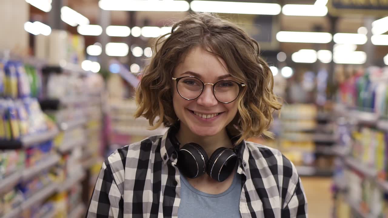 Portrait young woman stands in front of the camera and smiles in supermarket feel happy girl shopping face retail store. Pretty curly female customer in casual with headphones on neck with other customers on blurred background