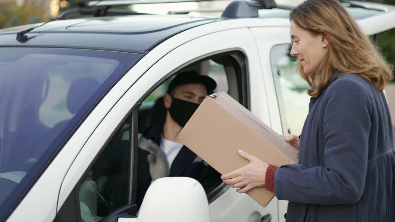 Delivery man wearing uniform, protection mask and gloves hand over a package to customer on the cargo van. Happy smiling female