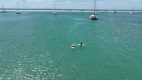 reverse flight over moored catamarans to reveal two men on surf boards